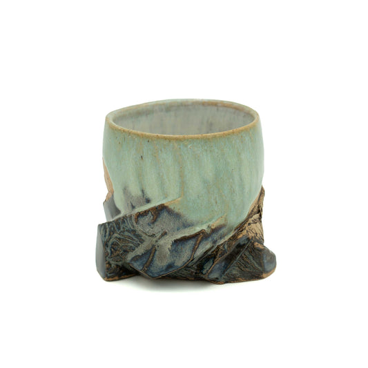 A handmade ceramic cup with a light blue-green glazed body and a dark brown textured, mountain ridges, shown on a plain white background.