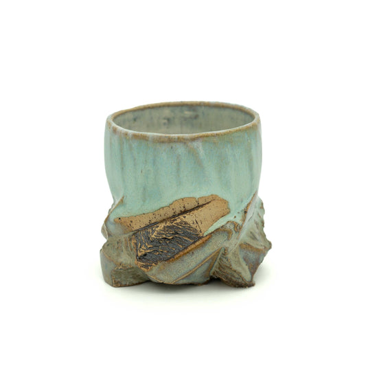 A small handmade ceramic cup with a light blue-green glaze and a rustic, textured base resembling natural stone or bark, shown on a plain white background.