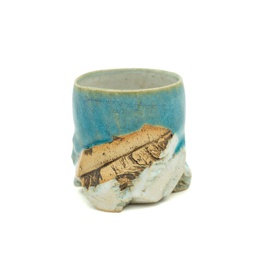 A handmade ceramic cup with a rustic, sculptural design, featuring a textured blue and turquoise glaze on the upper half, a natural wood slice embedded in the lower half, and a rough, off-white clay base. Photographed on a seamless white background.