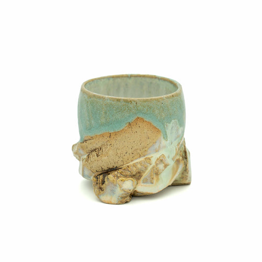 A small handmade ceramic cup with a textured, organic base resembling roots or leaves, featuring a light blue-green glaze and natural tan clay showing through, set against a plain white background.