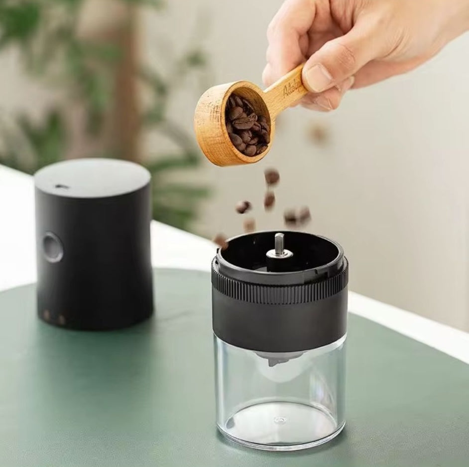 Person adding coffee beans from a wooden scoop into a clear coffee grinder on a green surface.
