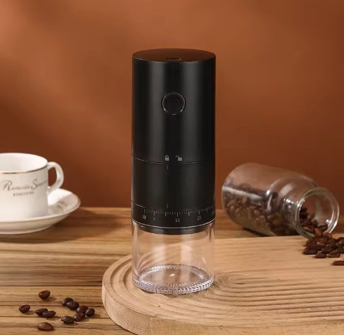 Black coffee grinder on a wooden surface with coffee beans and a cup in the background.